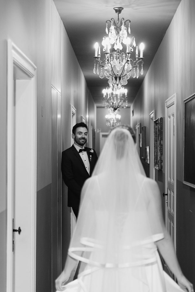 First Look eines eleganten Bräutigam im schwarzen Anzug Smoking und Braut im Hotelflur mit Kronleuchtern